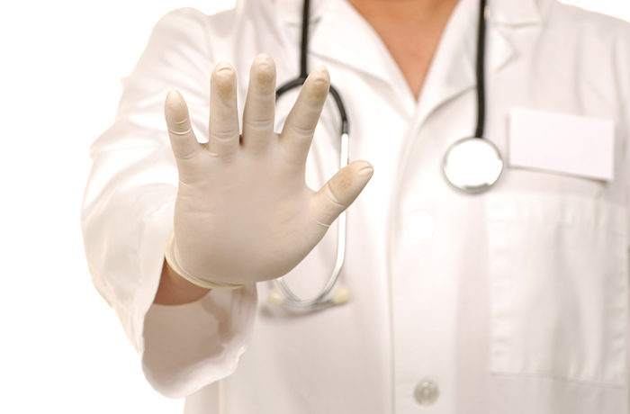 Frau mit Arztkittel, weißen Laborhandschuhen und Stethoskop um den Hals streckt die geöffnete rechte Hand aus zur Beruhigung.