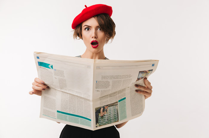 Junge Frau mit braunen Haaren und roter Baskenmütze vor grauem Hintergrund hält weit aufgeschlagene Zeitung vor sich und zeigt großes Erstaunen und Aufregung über den Inhalt.