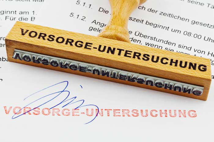 Holz-Stempel mit der Aufschrift Vorsorge-Untersuchung liegt auf einem gedruckten Papier mit rotem Stempelaufdruck und Unterschrift.
