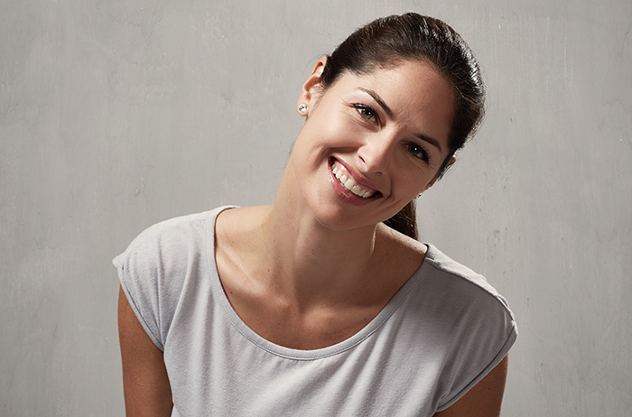 Freundlich lächelnde Frau mit langen braunen Haaren im grauen T-Shirt vor grauem Hintergrund schaut optimistich und überzeugend.