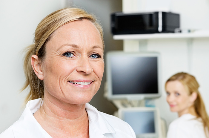 Blonde Ärztin strahlt zuversichtlich in die Kamera mit ihrer Kollegin im Hintergrund, die sich auch lächelnd zur Kamera dreht, beide in weißen Kitteln.