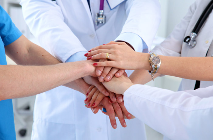 4 Ärzte im Kittel, teilweise mit Stethoskop, stehen zusammen und reichen sich die Hände und legen Sie in der Mitte übereinander.