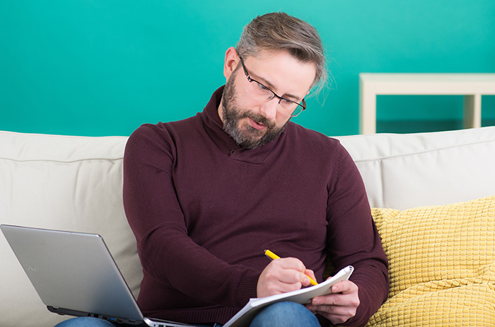 Mann mit kurzem Voll-Bart und Brille im auberginefarbenem Pullover sitz auf einer Couch mit Laptop und schreibt mit gelbem Kugelschreiber in einen Ringbuch-Block.