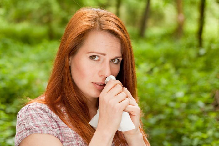Weinende junge Frau mit langen roten Haaren vor grünen Pflanzen und ein paar Baumstämmen hält weißes Taschentuch vor die Nase.
