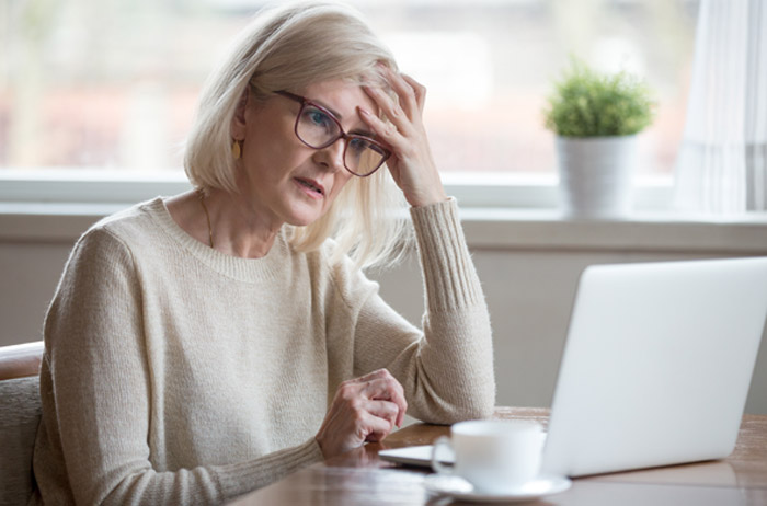 Ältere blonde Frau mit Brille, beigem Pullover und einer Hand an der Stirn sitzt bei einer Kafffeetasse vor ihrem weißen Laptop, im Hintergrund Fenster mit Grünpflanze im Blumentopf und geöffneter Gardine,  sie  schaut angestrengt überlegend auf den Laptop-Bildschirm.