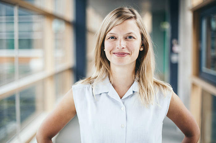 Junge blonde Frau mit weißer kurzärmliger Bluse steht in einem Gebäude, die Hände an den Hüften positioniert, und lächelt selbstbewusst in die Kamera.