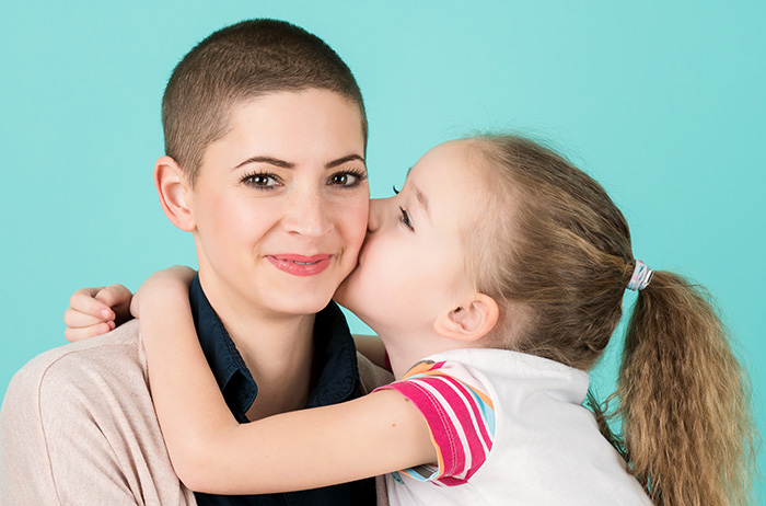 Kleines Mädchen mit Zopf küsst ihre lächelnde Mutter mit kurzen Haaren, die eine Krebspatientin ist/war, auf die Wange.