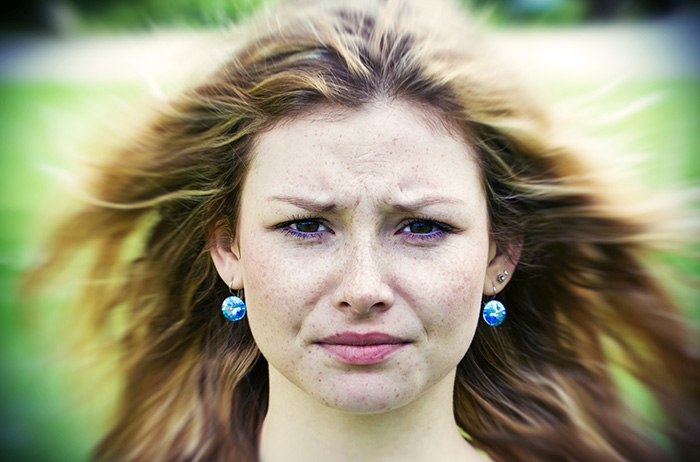 Junge Frau mit blonden Haaren, Sommersprossen und blauen Ohrringen schaut fragend und skeptisch, im Hintergrund grüne und verschwommene Landschaft.