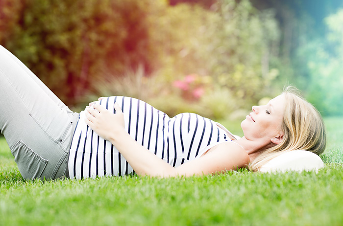 Blonde, schwangere Frau mit gestreiftem Oberteil liegt auf einer Wiese auf ihrem Rücken und blickt lächelnd auf ihren Bauch.