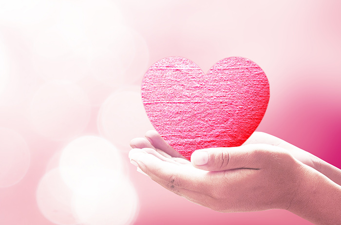 Zwei Hände, ausgestreckt und nach oben geöffnet tragen vorsichtig ein rosarotes Herz vor rosarotem Hintergrund