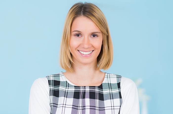 Junge blonde Frau im Kleid mit schwarz-weißem Karo-Muster und weißen Ärmeln vor hell-blauem Hintergrund lächelt überzeugt und selbstbewusst über die Leistungen im Service.