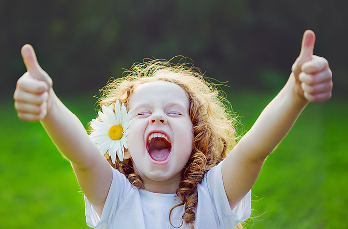 Auf einer Wiese streckt ein blondes junges Mädchen mit Blume im Haar beide Hände mit Daumen hoch und schreit vor Begeisterung.